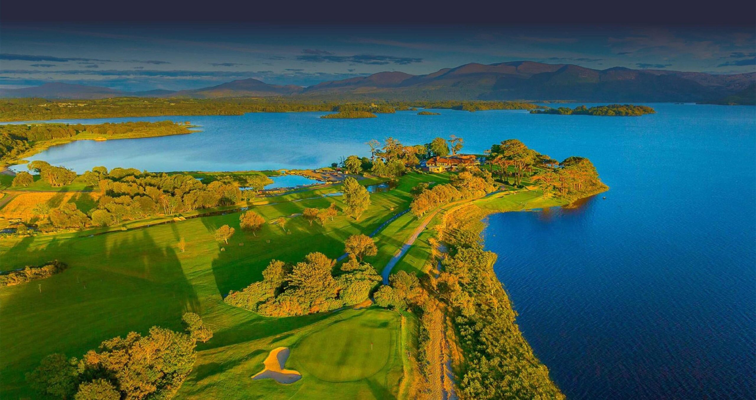 Killarney Golf Club in County Kerry, Ireland, featuring a scenic lakeside green, surrounded by lush greenery and majestic mountains in the background. 