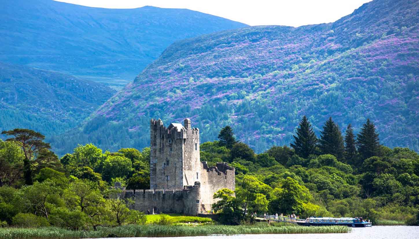 Ross Castle, a 15th-century tower house, sits on the shores of Lough Leane in Killarney National Park, County Kerry, Ireland, surrounded by lush greenery and majestic mountains. 