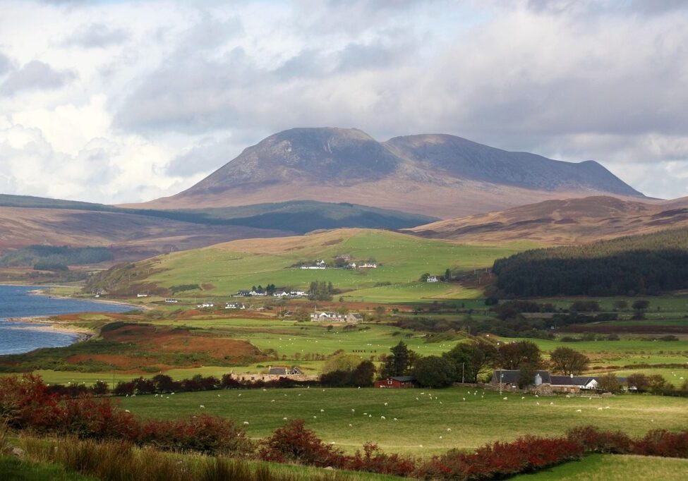 A scenic image of the isle of Arran with the mountains in the background, farmlands in the foreground, and the ocean to the left.